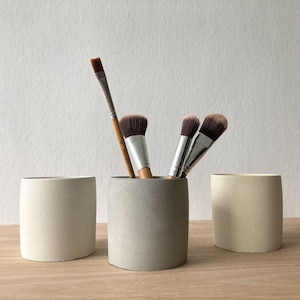 May include: Three white ceramic cylindrical containers with rounded edges, two are empty and one holds a set of makeup brushes, including a wooden handled brush and two black handled brushes.
