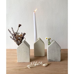 May include: Three small concrete house-shaped candle holders, two with dried flowers and one with a white candle. The candle holders are on a wooden surface.