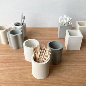 May include: A set of six small, concrete containers in various shapes and sizes. The containers are in shades of grey and white and are used to hold toothpicks, cotton buds, and other small items.