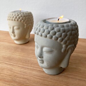May include: Two concrete candle holders shaped like Buddha heads. The candle holders are grey and white and have a textured surface. A tea light candle is burning in one of the holders.