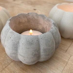 May include: Three grey concrete pumpkin-shaped candle holders. The centre holder has a white tea light candle burning inside.