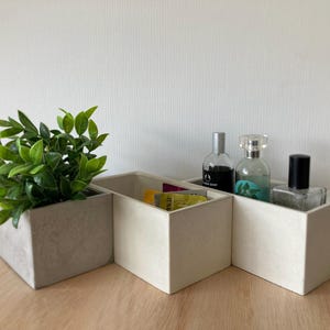 May include: A three-section organiser with a faux-concrete finish. One section holds a green plant, another contains small items, and the third holds perfume bottles. The organiser sits on a wooden surface.