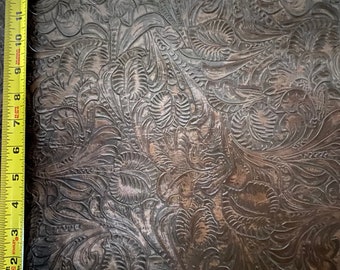 May include: Close-up of a dark brown leather surface with an embossed floral pattern. A yellow measuring tape is placed along the top edge of the surface.