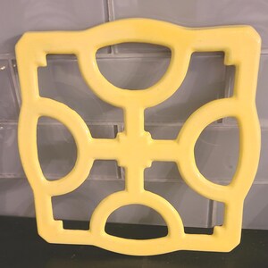 May include: A yellow, square trivet with a decorative openwork design. The trivet features rounded shapes and a cross-like pattern. This kitchen accessory is designed to protect surfaces from hot cookware.