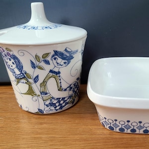 May include: A white ceramic sugar bowl with a lid and a small serving dish. The sugar bowl is decorated with blue and green illustrations of figures. The serving dish has a blue and white geometric pattern along the bottom edge.