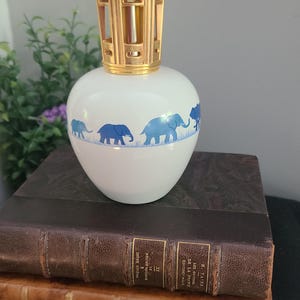 May include: A white ceramic fragrance lamp with a rounded body featuring a blue elephant parade pattern around the center. The lamp is topped with a gold-colored metal openwork cap. The design includes a horizontal blue line beneath the elephants.