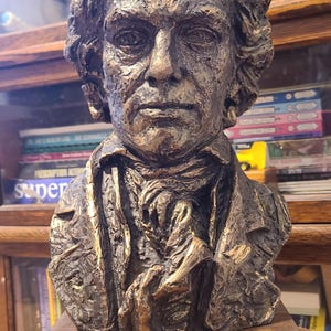 May include: A bronze-toned bust of a man with curly hair and a serious expression, mounted on a rectangular wooden base. The sculpture has detailed facial features and is set against a blurred background.