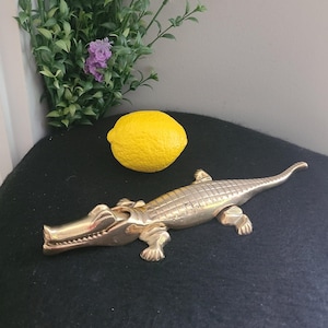 May include: A gold-coloured alligator nutcracker with a textured surface. The alligator is lying on a black surface with a yellow lemon beside it.