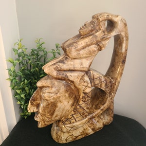 Peut inclure: Une sculpture en bois sculpté de trois visages abstraits, dans un schéma de couleurs brun et beige. La sculpture est faite d'une seule pièce de bois et a une finition lisse et polie.
