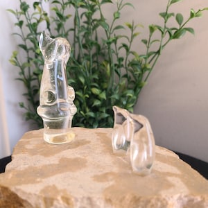 Op de afbeelding: Twee doorzichtige glazen beeldjes, een in de vorm van een hoge, slanke toren en de andere in de vorm van een pinguïn, worden tentoongesteld op een ruwe, lichtbruine steen.