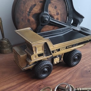 May include: A gold and black toy dump truck with black tires. The truck has the text "EUC R-50" and a logo on the side. A set of keys and a brass bell are in the foreground. A wooden wheel and a bell are in the background.