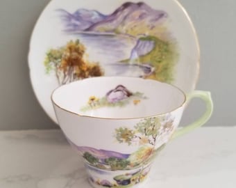 May include: A white porcelain teacup and saucer set featuring a hand-painted landscape scene with purple mountains, green trees, and a body of water. The teacup has a light green handle and a gold-trimmed rim, while the saucer has a scalloped edge.