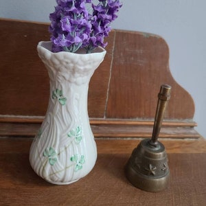 May include: A white ceramic vase with a textured design and green shamrock accents holds purple lavender flowers. A small brass bell with star cutouts sits to the right. The objects are on a wooden surface.