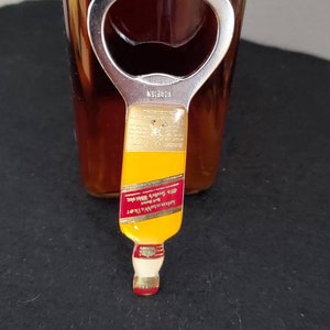 May include: A gold bottle opener shaped like a miniature bottle of Johnnie Walker Red Label Scotch Whisky. The bottle opener has a silver metal top with a bottle opener.