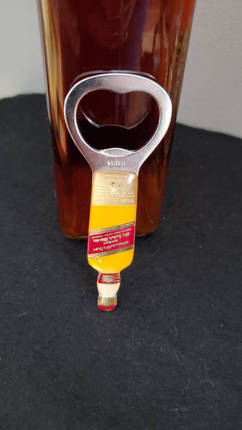 May include: A gold bottle opener shaped like a miniature bottle of Johnnie Walker Red Label Scotch Whisky. The bottle opener has a silver metal top with a bottle opener.