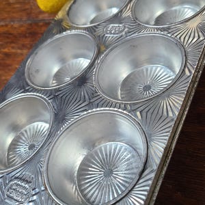 May include: A vintage, silver muffin tin with eight individual cups. The tin has a decorative, starburst pattern around each cup. A yellow lemon rests near the tin. The tin has the text "Ecko" and "Flint" embossed on it.