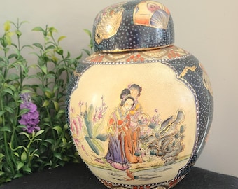 May include: A ceramic jar with a lid, decorated with a black and gold floral pattern. The jar features a scene of two women in traditional clothing standing in a garden.
