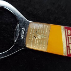May include: A silver bottle opener with a yellow and red John Walker & Sons Old Scotch Whisky label. The label has gold lettering and a small image of a man walking. The bottle opener is labeled "FOREIGN" on the side.