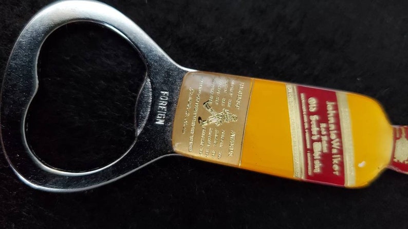 May include: A silver bottle opener with a yellow and red John Walker & Sons Old Scotch Whisky label. The label has gold lettering and a small image of a man walking. The bottle opener is labeled "FOREIGN" on the side.