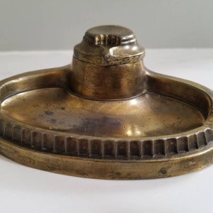 May include: A brass inkwell with a round, raised lid and a decorative, scalloped edge. The inkwell is oval shaped and has a shallow well for ink.