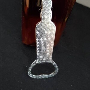 May include: Silver bottle opener with a textured surface and a round opening. The opener is designed to fit around the neck of a bottle.