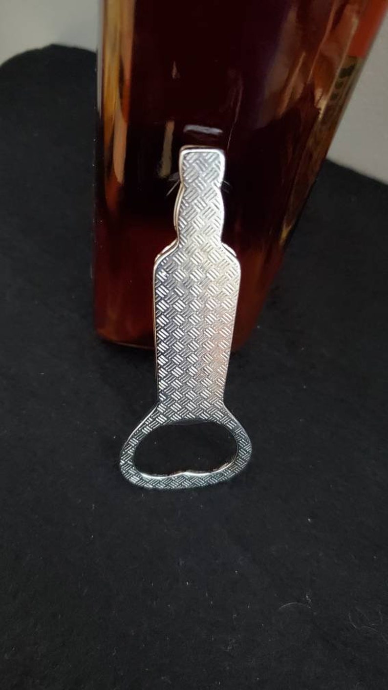 May include: Silver bottle opener with a textured surface and a round opening. The opener is designed to fit around the neck of a bottle.