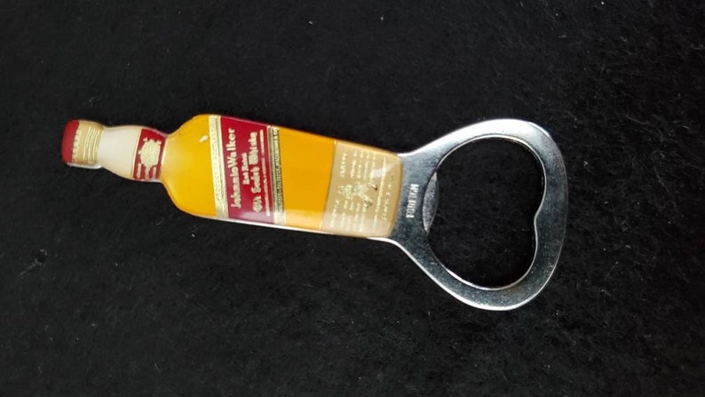 May include: A silver bottle opener with a yellow and red plastic handle. The handle features the text "Johnnie Walker" and "Old Scotch Whisky".