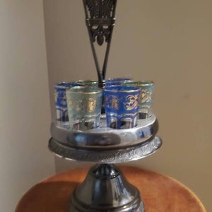 May include: A silver shot glass holder with a decorative, ornate design. The holder has six shot glasses with blue and gold designs. The holder is on a wooden table.