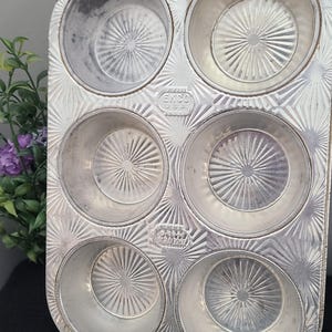 May include: A vintage silver muffin tin with six circular moulds. The tin features a starburst pattern and the text "EKCO U.S.A." is visible. There is a hole at the top for hanging. Suitable for baking muffins or cupcakes.