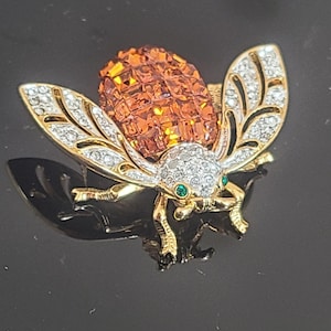 Op de afbeelding: Een gouden broche in de vorm van een kever met oranje kristallen vleugels en groene kristallen ogen.