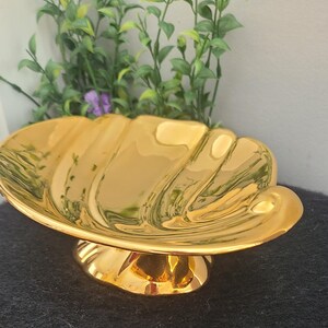 May include: A gold-colored, shell-shaped decorative dish with a pedestal base. The dish has a glossy finish and is set against a dark background with green foliage in the background. The dish is likely used for decorative purposes.