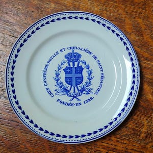May include: A white ceramic plate featuring a blue decorative border of leaves and a central blue emblem. The emblem displays a crown, a shield with a cross, and text reading CHEF CONFRÉRIE ROYALE ET CHEVALIÈRE DE SAINT SÉBASTIEN GAND FONDÉE EN 1322.