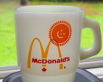 May include: White ceramic mug with a red and yellow McDonald's logo, a yellow sun with a red face, and the text "McDonald's" in red. The mug has a Canadian maple leaf.