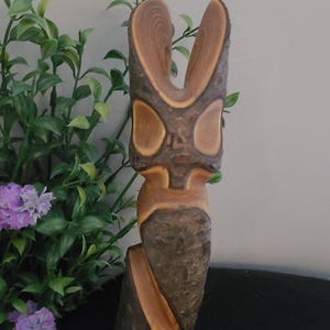 May include: A hand-carved wooden sculpture with a natural, rustic aesthetic. The sculpture features a stylized, abstract form with a central opening and rounded features. The wood's natural grain and colour variations, from light brown to dark brown, are visible.