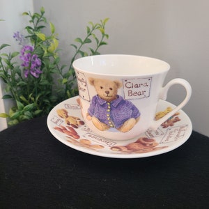 May include: A white teacup and saucer with a teddy bear design. The teacup features a brown teddy bear wearing a purple sweater and a sign that reads "Clara Bear". The saucer has a variety of brown and white teddy bears and the text "Clarence".