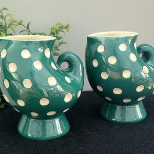 May include: Two green ceramic pitchers with white polka dots. Each pitcher has a unique handle and sits on a pedestal base. The pitchers are displayed on a dark surface with greenery in the background.