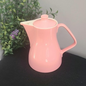 Può includere: Una teiera in ceramica rosa con coperchio e manico. La teiera ha un beccuccio curvo e un interno bianco. Lo sfondo comprende fogliame verde e un muro grigio. La teiera è di colore rosa chiaro.