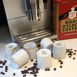Op de afbeelding: Een zilveren Jura ENA koffiemachine staat naast een rode Eight O'Clock koffiebus. Vijf witte porseleinen mokken met gouden randen en oren liggen verspreid op het oppervlak, omringd door donkere koffiebonen. De mokken zijn voorzien van Limoges-merktekens.