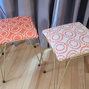 Könnte beinhalten: Zwei quadratische Hocker mit goldenen Haarnadelbeinen aus Metall. Die Sitze sind mit einem weißen Stoff mit einem sich wiederholenden Muster aus orangefarbenen Kreisen gepolstert. Ein Hocker hat einen orangefarbenen Hintergrund, der andere einen weißen Hintergrund.