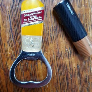 May include: A silver bottle opener with a yellow and red Johnie Walker Red Label Scotch Whisky bottle design. The bottle opener has the word "FOREIGN" printed on it.