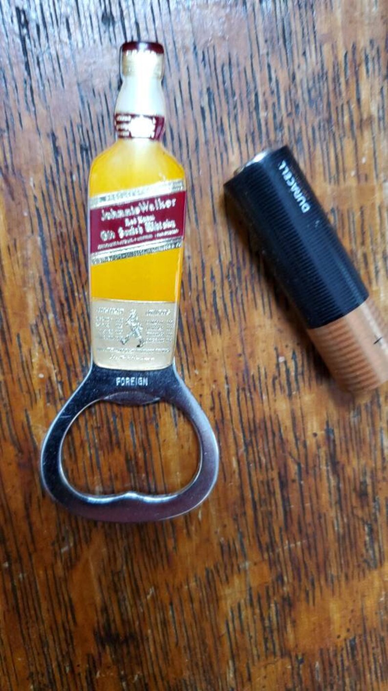 May include: A silver bottle opener with a yellow and red Johnie Walker Red Label Scotch Whisky bottle design. The bottle opener has the word "FOREIGN" printed on it.