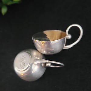 May include: Two silver cups with a gold interior. The cups have a rounded shape with a handle on one side. The bottom of one cup has the inscription "Sheffield England" on it.