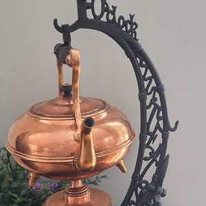 Op de afbeelding: Een koperen ketel met een tuit en handvat, opgehangen aan een zwarte smeedijzeren standaard. De standaard heeft decoratief krulwerk en het woord "O'clock" bovenaan. De ketel staat op een ronde basis met drie kleine voetjes.