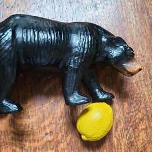 May include: A black bear figurine with a brown snout stands next to a yellow lemon on a wooden surface. The bear is carved with visible texture and detail. The lemon is a bright, contrasting colour.