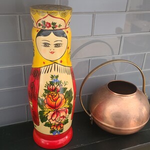 Könnte beinhalten: Eine handbemalte Matrjoschka-Puppe aus Holz mit gelbem Kopftuch und floralem Design. Die Puppe steht neben einer Gießkanne aus Kupfer mit gebogenem Griff. Die Puppe ist etwa 25 cm hoch.