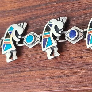 May include: Silver bracelet featuring Kokopelli figures with turquoise, lapis, and coral accents. The bracelet has a series of Kokopelli figures alternating with eye-shaped links. The bracelet is set against a dark wood grain background.