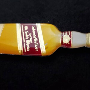 May include: A silver bottle opener shaped like a John Walker & Sons Old Scotch Whisky bottle. The bottle opener has a red, gold, and white label with the text "Johnnie Walker Old Scotch Whisky" printed on it.