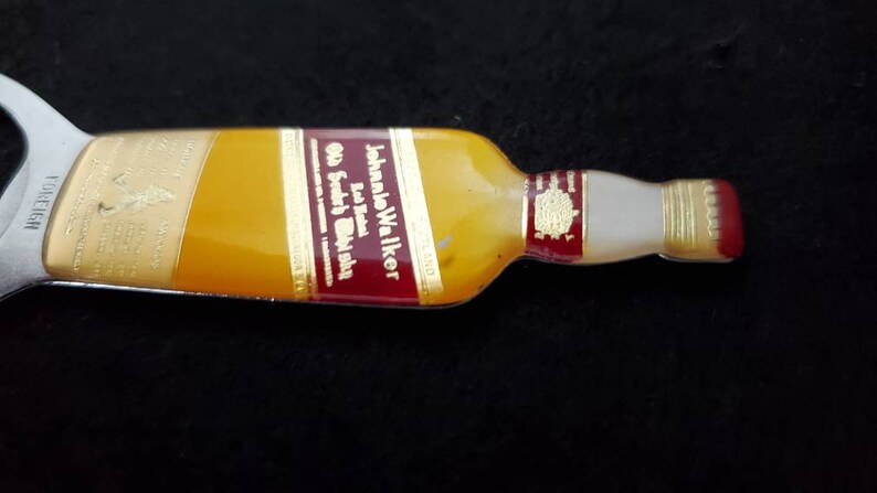 May include: A silver bottle opener shaped like a John Walker & Sons Old Scotch Whisky bottle. The bottle opener has a red, gold, and white label with the text "Johnnie Walker Old Scotch Whisky" printed on it.