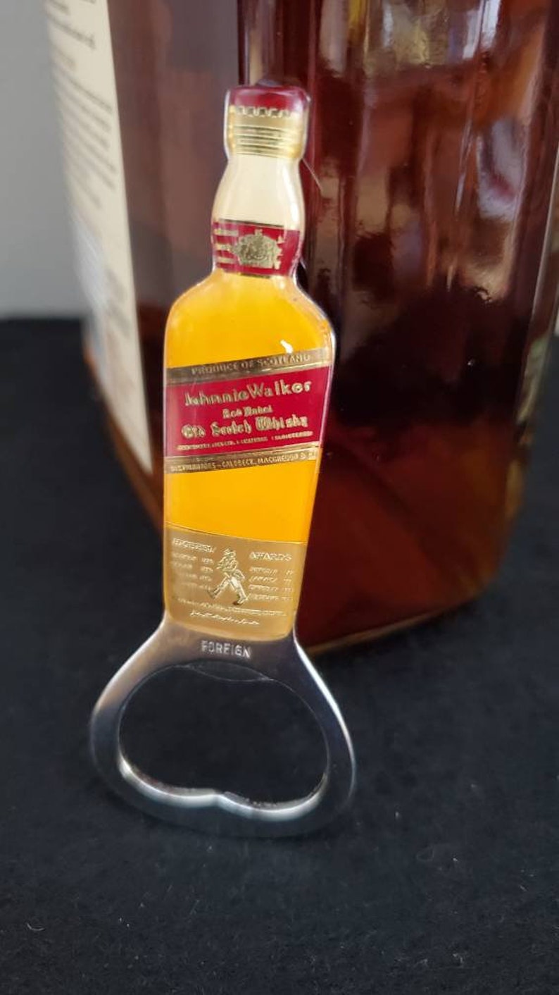 May include: A gold bottle opener with a red and gold Johnnie Walker Red Label Old Scotch Whisky label. The bottle opener has the words "Foreign" and "Avatars" printed on it.