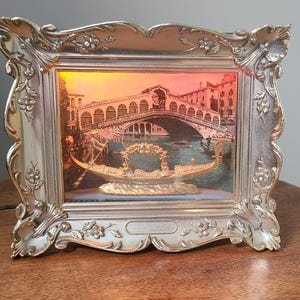 Può includere: Opera d'arte incorniciata color argento con una scena veneziana con il Ponte di Rialto e una gondola. La cornice presenta dettagli floreali ornati. L'opera d'arte ha un bagliore caldo, simile al tramonto, creando un'atmosfera romantica.